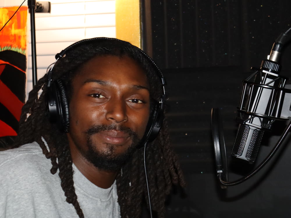 A vocalist wearing headphones smiles beside a large condenser microphone and pop filter inside the vocal booth.