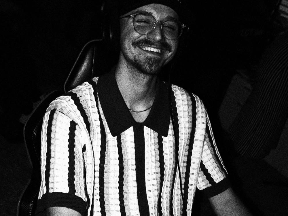 A producer in a striped shirt, ball cap and glasses laughs at the studio desk, headphones on, in high-contrast black and white.