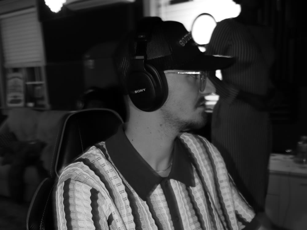 An engineer in headphones and a cap works at the console in profile while a vocalist stands in the background, in black and white.