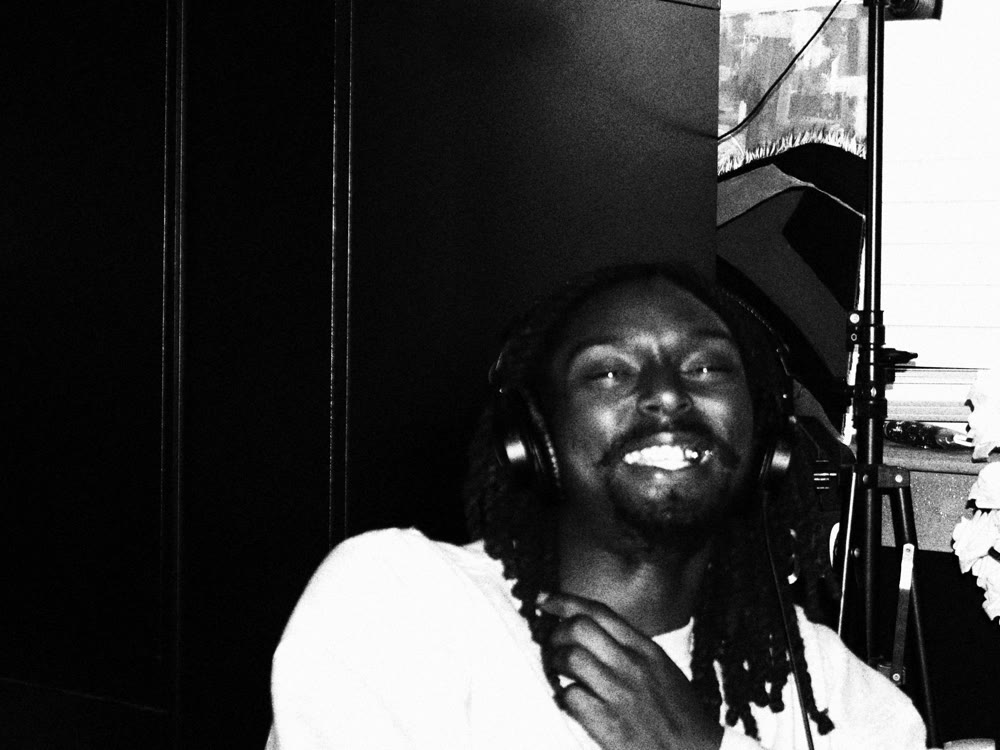 A musician wearing headphones laughs beside a mic stand during a recording session, in high-contrast black and white.
